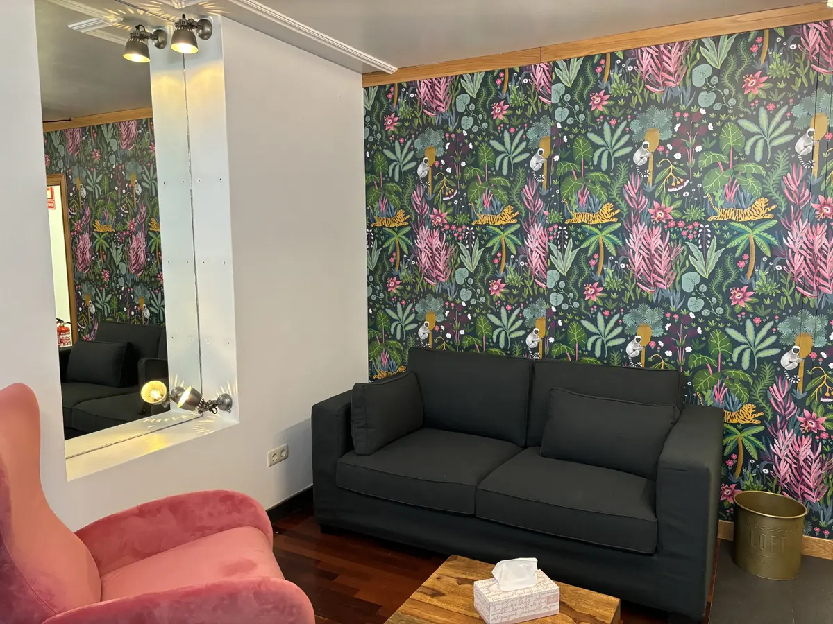 Zona de conversación de la consulta: sofá gris oscuro, butaca de terciopelo rosa y pared con papel pintado botánico